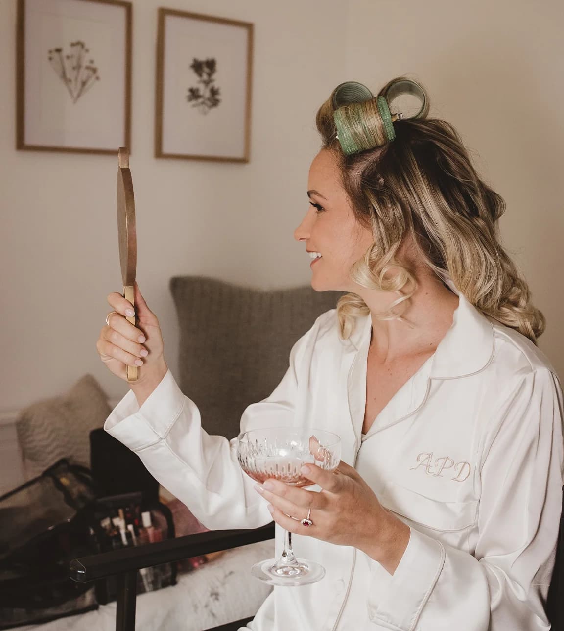 Bride getting hair and makeup done with curlers in and wearing a monogrammed pyjama set while holding a glass of champagne