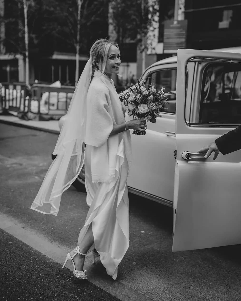 Smiling bride wearing a veil and holding bouquet getting into a cream London taxi